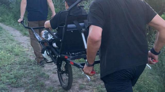 tres personas en un camino mucha naturaleza alrededor, uno de ellos es llevado en una silla adpatada para senderismo accesible