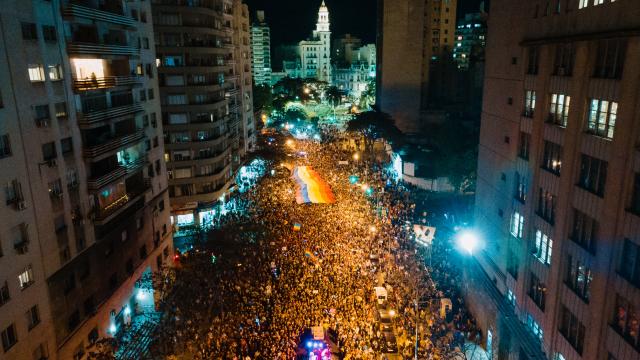 "Vista aérea nocturna de la Marcha de la Diversidad en Montevideo, con una multitud llenando la Avenida 18 de Julio y una gran bandera arcoíris extendida en el centro, iluminada por las luces de la ciudad y con el Palacio Salvo visible al fondo.