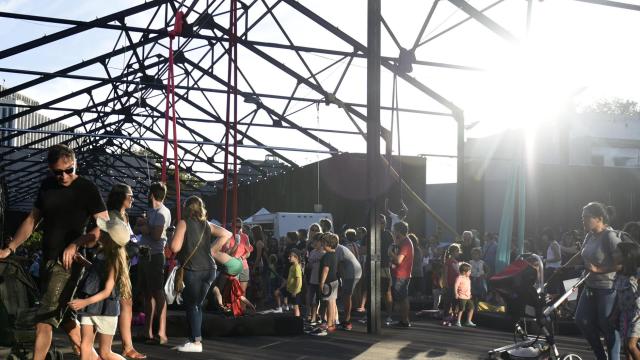 Espacio abierto con estructura metálica de techo sin cubrir, lleno de familias y niños disfrutando de actividades recreativas al atardecer. Algunas personas caminan, otras empujan cochecitos y varios niños juegan cerca de columpios y telas acrobáticas, con la luz del sol entrando desde el fondo.