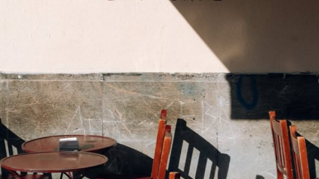 Mesas y sillas de madera al aire libre en la vereda de un bar; en la pared blanca se lee el cartel “Santa Catalina Bar·Café”.