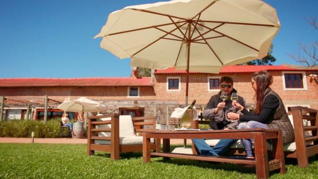 Pareja sentada al aire libre en sillones de madera con cojines, bajo un gran paraguas blanco. Brindan con copas de vino frente a la fachada de ladrillo de la Bodega Valdi, en un día soleado.