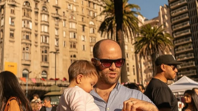 Hombre con gafas de sol sostiene en brazos a una niña pequeña mientras come en la Plaza Independencia de Montevideo. De fondo se ven el Palacio Salvo, palmeras y varias personas disfrutando de una jornada al aire libre.