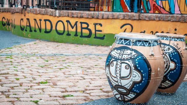 Tambores de candombe pintados con rostros afrodescendientes sobre un empedrado, junto a un mural que dice “Candombe”.