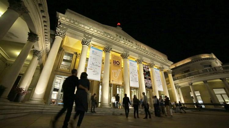Explanada con público llegando al espectáculo. Fachada principal del teatro iluminada. Se destaca sus columnas y su escalinata así como el nombre iluminado y la farola roja. 