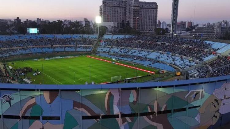 Foto del estadio, se ve la cancha con pasto verde, público en las gradas y las paredes exteriores del estadio pintadas. También se ven las torres con las luces iluminadas y el atardecer. 