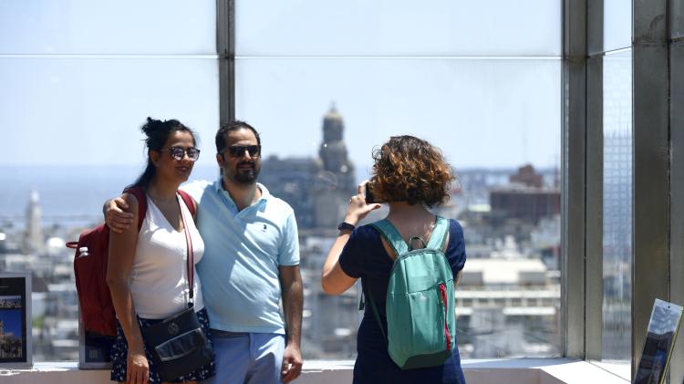 Dos personas sacándose una foto. Mirador vidriado. Al fondo edificios varios. 
