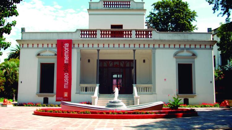 Fachada princial de la casa del museo pintada de blanco, escalinata central para el ingreso, bandera roja con nombre del museo a lo largo de las paredes. Delante de la fachada,fuente circular blanca con agua. 
