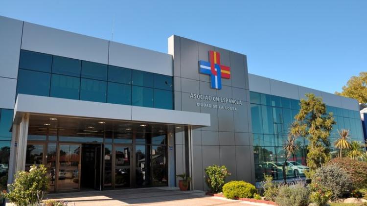 Una de las sedes del hospital en un día soleado. Construcción moderna de color gris con mucho vidrio templado. Logo de la empresa en la fachada en color azul y rojo. Pasto y arboles al frente. 