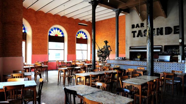 Salón principal con varias mesas y sillas iluminada por la luz del sol que refleja en los vitrales de colores. Encontramos también una estatua de Don Quijote y el horno de pizza con el nombre Tasende arriba y su barra de azulejos antiguos. También tiene paredes de ladrillos. 