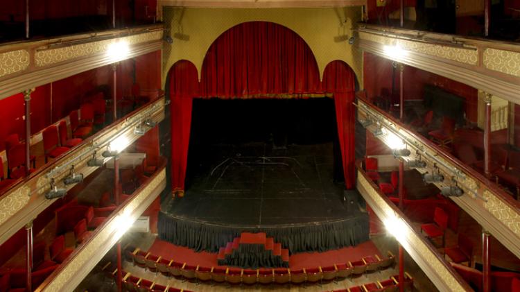 Panorámica del teatro a la italiana en forma de herradura. Al fondo escenario principal con telones rojos. A los costados palcos y butacas rojas en diferentes pisos. Platea debajo. 