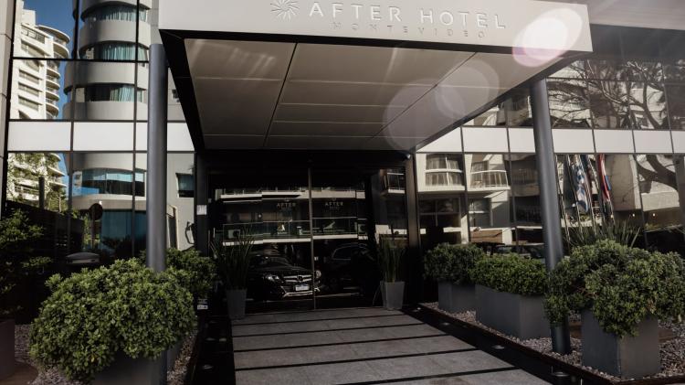 Entrada moderna al hotel. Hay una puerta de vidrio corrediza. Hay plantas y arbustos en el ingreso. 