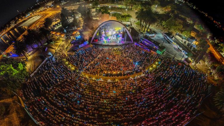 Vista aérea del Teatro de Verano repleto de personas observando un espectáculo que se está llevando a cabo en su escenario. La platea es abierta y el escenario tiene un techo en forma de una gran bóveda ovalada.