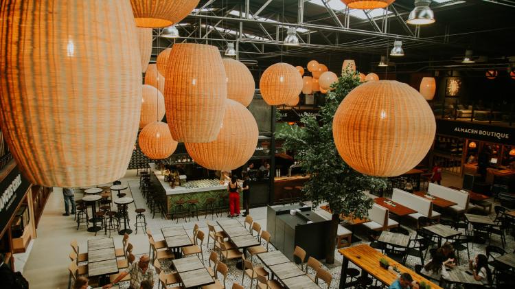 Foto desde arriba de interior del mercado destancando sus lamparas tipo mimbre ovales y redondas, gran plaza de comidas con mesas del mismo color en madera clara y barras de locales gastronomicos alrededor