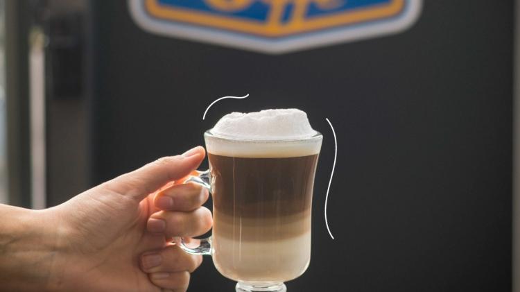 Mano sosteniendo un vaso de capuchino al fondo el cartel de la marca