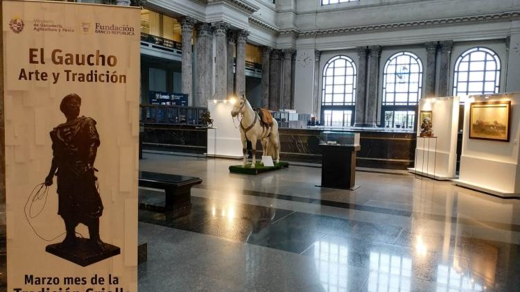 Vemos un hall, en primer plano un banner con la inscripción "El Gaucho. Arte y tradición. Marzo, el mes de la tradición criolla. Más atrás, un banco, una maqueta de un caballo, dos paneles blancos con cuadro y una caja con vidrio transparente para exhibición de objetos. 